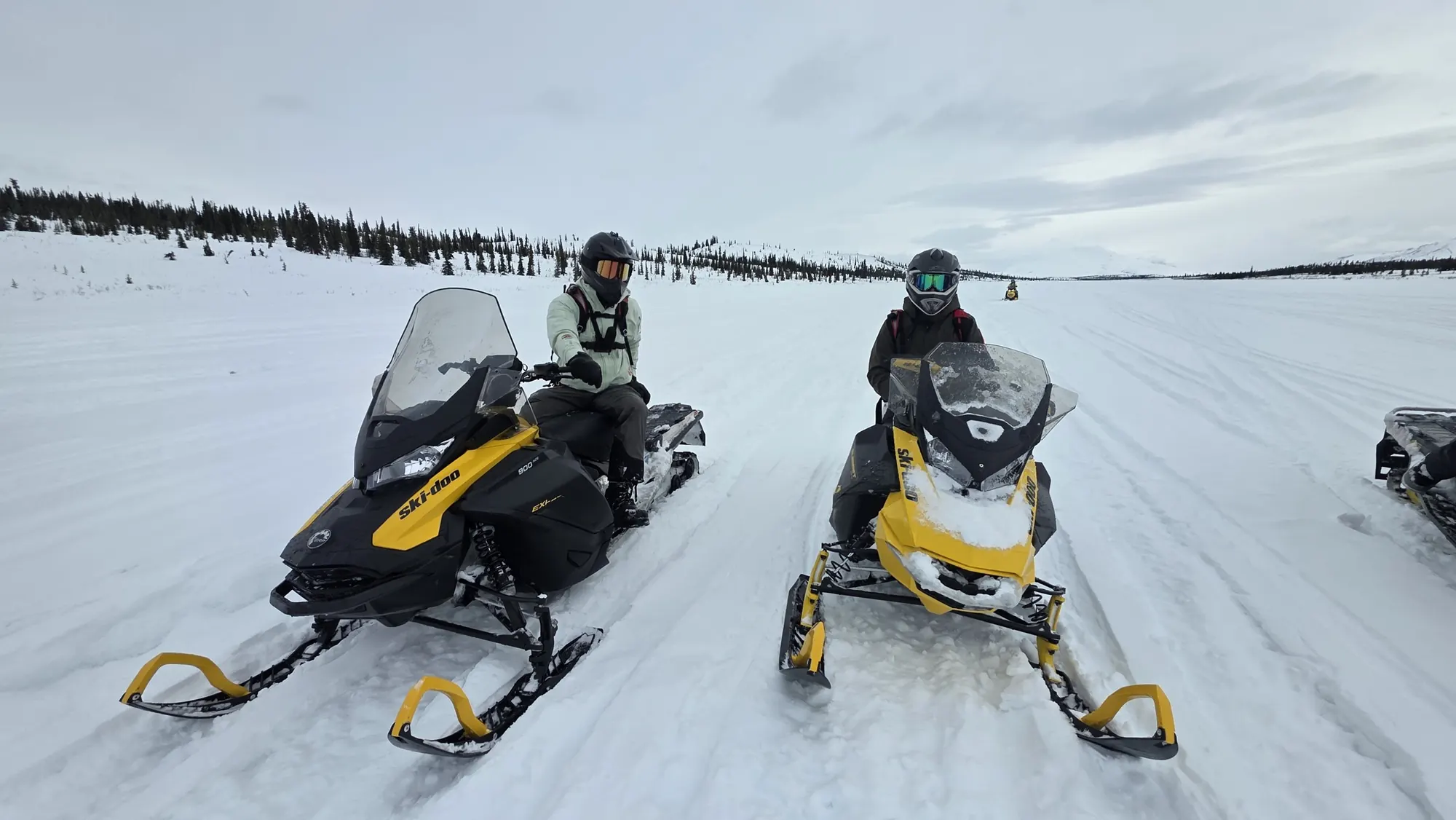 Two riders on yellow snowmobiles on a wide, snow-covered plateau under a grey sky